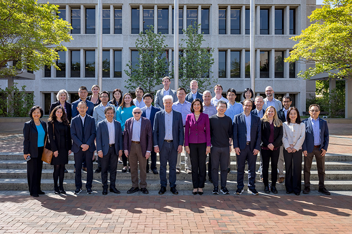 Group photo A large group of smiling men and women wearing business casual clothing pose outdoors in front of a campus building.