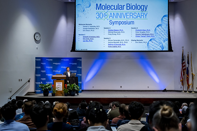 Srvivastata Overview Person at podium on-stage, speaks to an audience, in front of a screen showing the Molecular Biology 30th Anniversary Symposium agenda.