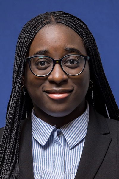 Zoe Osei-Appiah headshot; Black woman with long braids, glasses wearing dark jacket over striped blue shirt