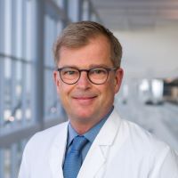 Markus Velten, M.D., white man, grey hair, black rimmed glasses, smiling, posed in front of blurred hospital background