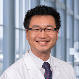 Dr. Jeffrey Chen wearing glasses, a white shirt with a patterned tie, and a white lab coat standing in the Clements University Hospital lobby