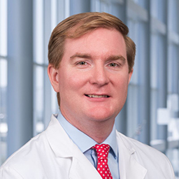 Dr. Ben Nelson wearing a light blue shirt with a patterend red tie and a white lab coat standing in the Clements University Hospital lobby