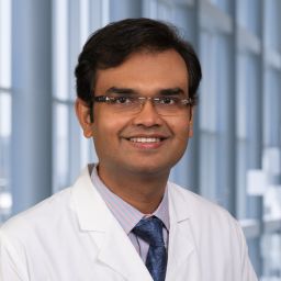Smiling man with dark hair and glasses, wearing a white lab coat.
