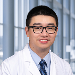 Dr. Jianyi Yin wearining glasses, a light blue shirt with blue stipped necktie and standing in the Clements University Hospital lobby