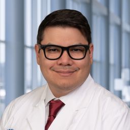 Dr. Julio Alvarenga Thiebaud wearing glasses and a white shirt with a red tie and a white lab coat standing in the Clements University Hospital lobby
