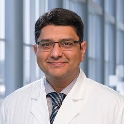 Dr. Farrukh Awan wearing a grey shirt with a blue stiped tie and a white lab coat standing in the Clements University Hospital lobby