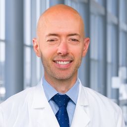 Dr. James Brugarolas wearing a light blue shirt with a dark blue tie and a white lab coat standing in the Clements University Hospital lobby