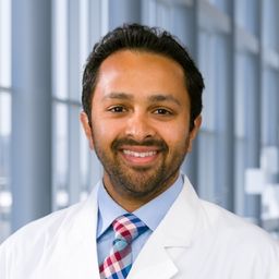 Dr. Sanjay Chandrasekaran wearing a light blue shirt with red, white and blue checkered tie a and a white lab coat standing in the Clements University Hospital lobby