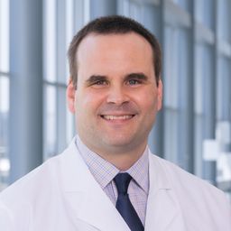 Dr. Kevin Courtney wearing a light grey shirt with a dark tie and a white lab coat standing in the Clements University Hospital lobby