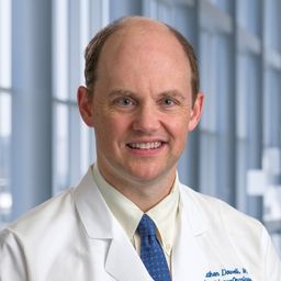 Dr. Jonathan Dowell wearing a beige shirt with a blue tie and a white lab coat standing in the Clements University Hospital lobby