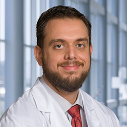 Dr. Ibrahim Ibrahim wearing a light grey shirt with a red tie and a white lab coat standing in the Clements University Hospital lobby