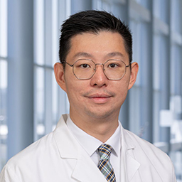Dr. Charles Jiang wearing a white shirt with a plaid tie and a white lab coat standing in the Clements University Hospital lobby