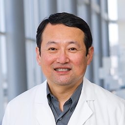 Dr. James Kim wearing a dark grey shirt and a white lab coat standing in the Clements University Hospital lobby