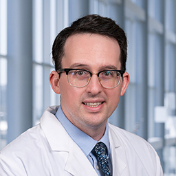 Dr. Jake Lichterman wearing a light blue shirt with a blue patterned tie and a white lab coat standing in the Clements University Hospital lobby