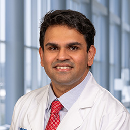 Dr. Nikhil Madhusudhan wearing a light blue shirt with a red patterned tie and a white lab coat standing in the Clements University Hospital lobby
