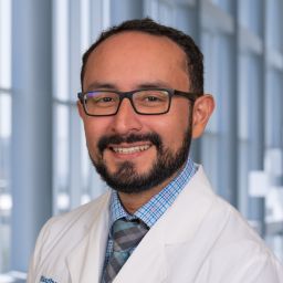 Dr. Alejandro Marinos Velarde wearing a light blue plaid shirt and a white lab coat standing in the Clements University Hospital lobby