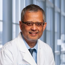 Dr. Syed Mazher wearing a light blue shirt with a dark blue tie and a white lab coat standing in the Clements University Hospital lobby