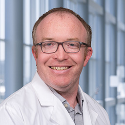 Dr. Patrick McCreesh wearing a grey shirt and a white lab coat standing in the Clements University Hospital lobby