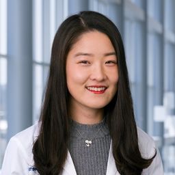 Dr. Qian Qin wearing a grey shirt and a white lab coat standing in the Clements University Hospital lobby