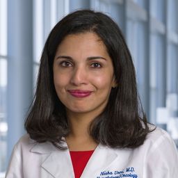 Dr. Nisha Unni wearing a red shirt and a white lab coat standing in the Clements University Hospital lobby