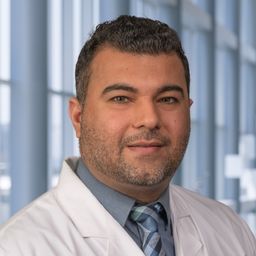 Dr. Nilesh Verma wearing a dark grey shirt with a plaid tie and a white lab coat standing in the Clements University Hospital lobby