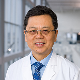 Dr. Jue Wang wearing a blue shirt with a blue patterned tieand a white lab coat standing in the Clements University Hospital lobby