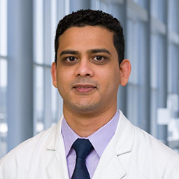 Dr. Mir Alikhan wearing a lavender shirt with a dark blue tie and a white lab coat standing in the Clements University Hospital lobby