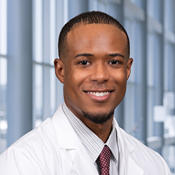 Dr. Kalonji Cole wearing g a light striped with a maroon patterned tie and a white lab coat standing in the Clements University Hospital lobby