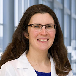 Dr. Antonia Dalton wearing glasses, a dark shirt, and a white lab coat standing in the Clements University Hospital lobby