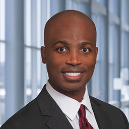 Dr. Abram Graham wearing a white shirt with a maroon tiew, and a dark suit coat standing in the Clements University Hospital lobby