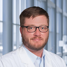 Dr. Patrick Green wearing glasses, a light blue shirt, and a white lab coat standing in the Clements University Hospital lobby