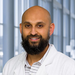 Dr. Athar Haq with a beard wearing a striped shirt, and a white lab coat standing in the Clements University Hospital lobby