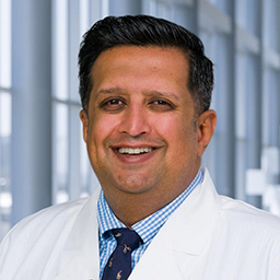Dr. Neeraj Kalra wearing a blue and white checkard shirt with a dark tie and a white lab coat standing in the Clements University Hospital lobby