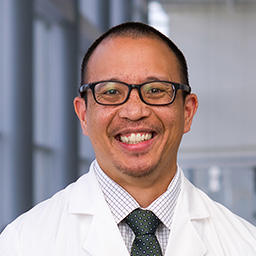 Dr. Noah A. Liwag wearing glasses, a light checkered shirt with dark tie, and a white lab coat standing in the Clements University Hospital lobby