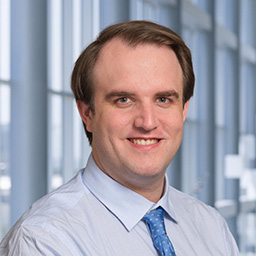Dr. James Lyons wearing a light blue shirt with a blue patterened tie, and a white lab coat standing in the Clements University Hospital lobby