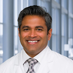 Dr. Satyam Nayak wearing a white shirt with a neutral plaid tie and a white lab coat standing in the Clements University Hospital lobby