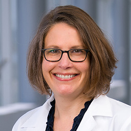 Dr. Christiana Renner wearing glasses, a dark ruffled shirt, and a white lab coat standing in the Clements University Hospital lobby