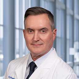 Dr. Bruce Stewart wearing a with shirt with a dark tie and a white lab coat standing in the Clements University Hospital lobby