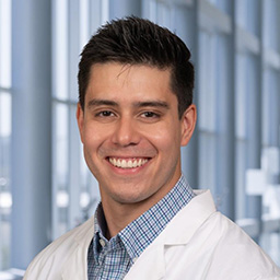 Dr. Jose Vargas Alvarado wearing a blue plaid shirt, and a white lab coat standing in the Clements University Hospital lobby