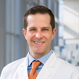 Dr. Paul Broker wearing a blue shirt with an orange tie, and a white lab coat standing in the Clements University Hospital lobby