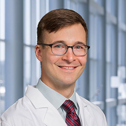 Dr. Daniel Driver wearing alight blue shirt with a red stiped tie and a white lab coat standing in the Clements University Hospital lobby