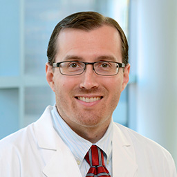 Dr Jason Newman wearing a light striped shirt with a red striped tie and a white lab coat standing in the Clements University Hospital lobby