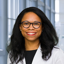 Dr. Nicole Oakman wearing glasses, a black shirt, and a white lab coat standing in the Clements University Hospital lobby