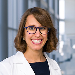Dr. Courtney Shipman-Brownlee wearing glasses, a black shirt, and a white lab coat standing in the Clements University Hospital lobby