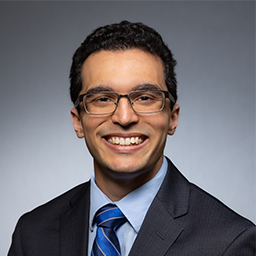 Christopher Joshi wearing glasses, a light blue shirt with a stripped blue necktie and dark grey jacket