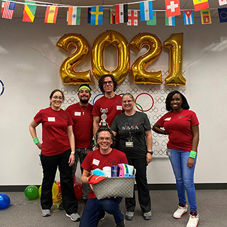 Team of approximately six people pose in front of olympic banner and balloons that spell out 2021