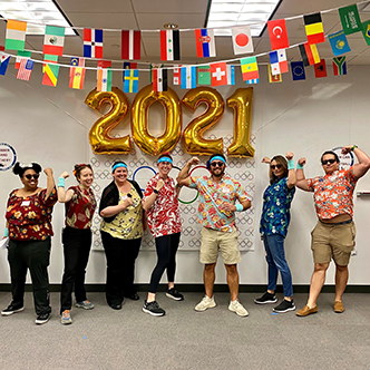 Team of approximately six people pose in front of olympic banner and balloons that spell out 2021