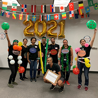 Team of approximately six people pose in front of olympic banner and balloons that spell out 2021