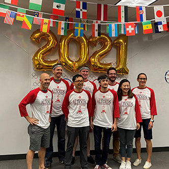 Team of approximately six people pose in front of olympic banner and balloons that spell out 2021