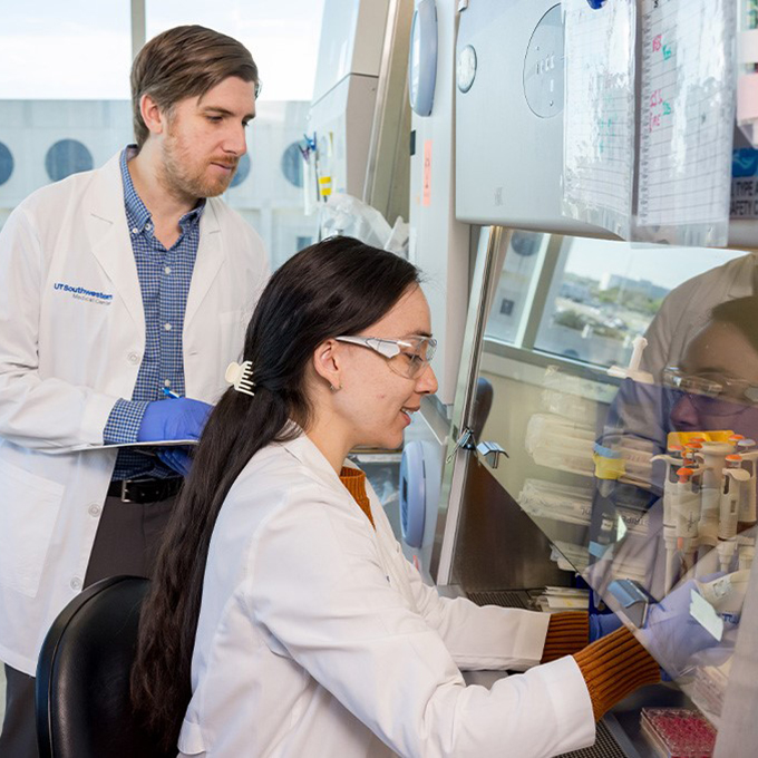 Male wearing lab coat is standing and taking notes aside a female wearing a lab coat sitting and performing a task at a desk in a lab